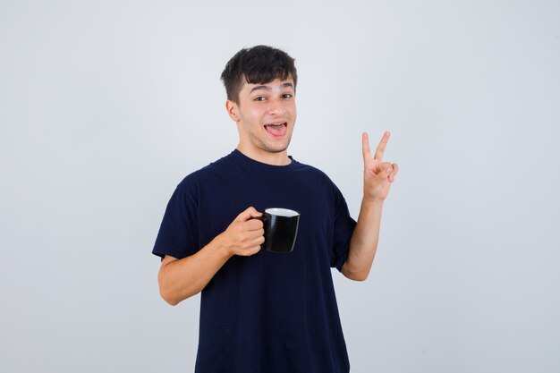 Młody człowiek w czarnej koszulce, trzymając filiżankę herbaty, pokazując znak V i patrząc szczęśliwy, widok z przodu.