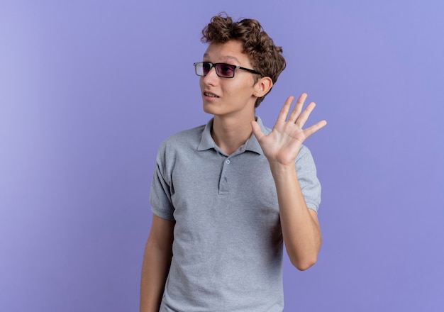 Młody człowiek w czarnych okularach na sobie szarą koszulkę polo, patrząc na bok uśmiechnięty macha ręką stojącą na niebieskiej ścianie