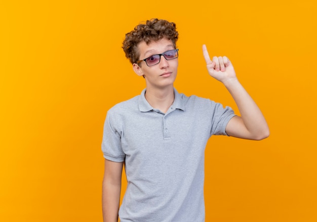 Młody człowiek w czarnych okularach na sobie szarą koszulkę polo szczęśliwy i pozytywny pokazuje palec wskazujący o nowym pomyśle na pomarańczowo
