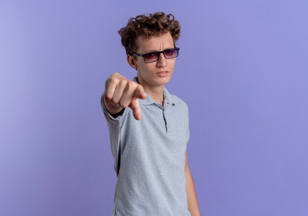 Młody człowiek w czarnych okularach na sobie szarą koszulkę polo, wskazując palcem wskazującym na aparat niezadowolony na niebiesko