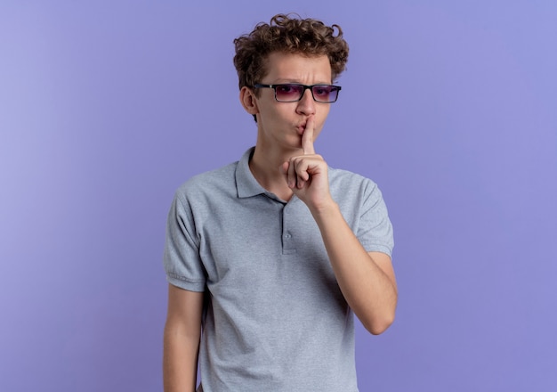 Młody człowiek w czarnych okularach na sobie szarą koszulkę polo z poważną twarzą robi gest ciszy z palcem na ustach stojąc na niebieskiej ścianie