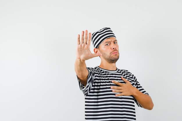 Młody człowiek w kapeluszu t-shirt w paski pokazuje gest stopu z ręką na klatce piersiowej i wygląda na zirytowanego