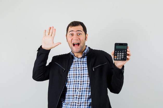 Młody człowiek w koszuli, kurtka trzymając kalkulator, pokazując dłoń i wyglądający na szczęśliwego
