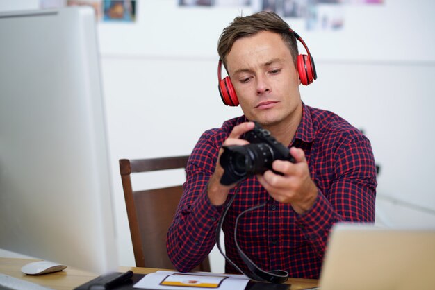 Młody człowiek w kraciastej koszuli i słuchawki siedzi przy biurku, trzymając aparat cyfrowy i patrząc na zdjęcia