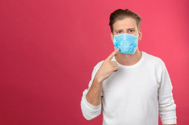 Młody człowiek w medycznej masce ochronnej patrząc na kamerę, wskazując palcem na maskę medyczną, musisz nosić maskę, aby uniknąć zachorowania na izolowanym różowym tle