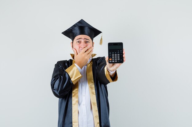 Młody człowiek w mundurze absolwenta trzymając kalkulator i patrząc niespokojnie, widok z przodu.