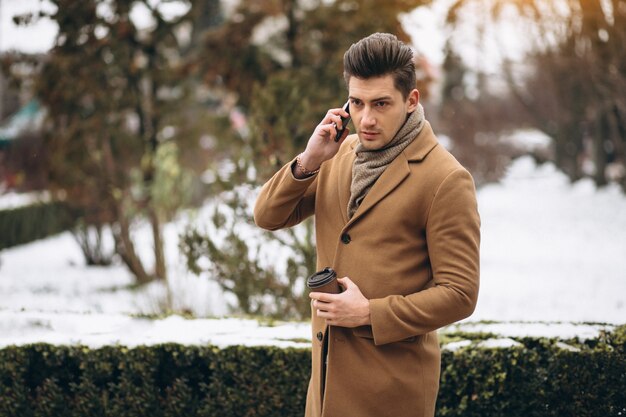 Młody człowiek w płaszczu poza rozmawia przez telefon i picia kawy