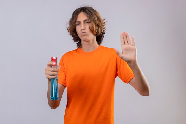 Młody człowiek w pomarańczowej koszulce trzymający spray do czyszczenia robiący znak stopu z gestem obrony dłoni patrząc na kamerę z marszczoną twarzą stojącą na białym tle