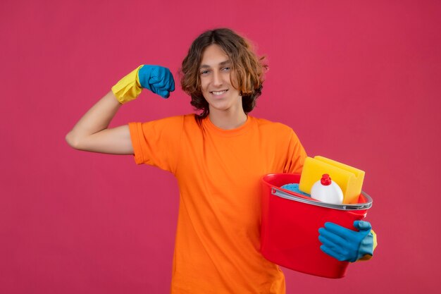 Młody człowiek w pomarańczowej koszulce w gumowych rękawiczkach trzyma wiadro z narzędziami do czyszczenia, podnosząc pięść, ciesząc się swoim sukcesem i zwycięstwem szczęśliwy i pozytywny uśmiechnięty stojący na różowym tle