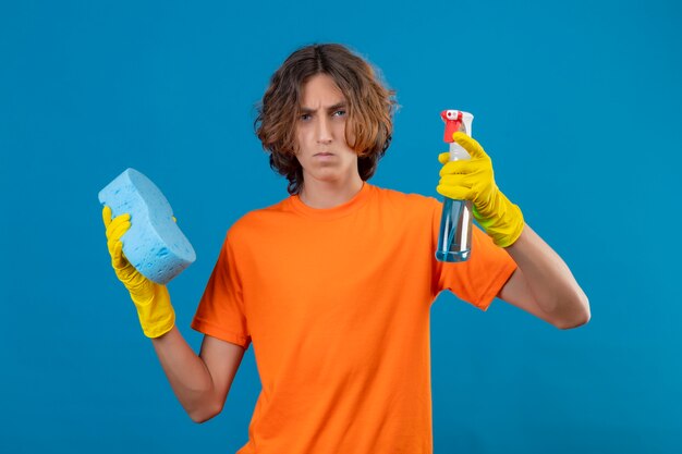 Młody człowiek w pomarańczowej koszulce w gumowych rękawiczkach, trzymając gąbkę i spray do czyszczenia patrząc na kamery z poważną twarzą stojącą na niebieskim tle