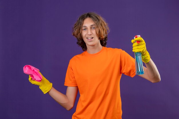 Młody człowiek w pomarańczowej koszulce w gumowych rękawiczkach, trzymając spray do czyszczenia i dywan, patrząc na kamery z pewnym uśmiechem stojącym na fioletowym tle