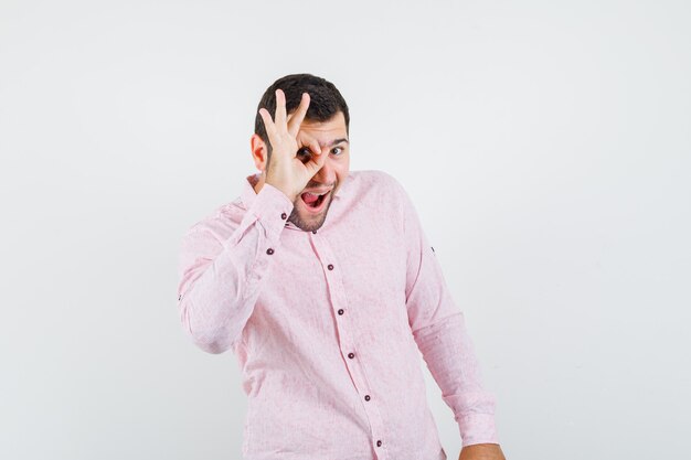Młody człowiek w różowej koszuli pokazuje znak ok na oko i wygląda śmiesznie