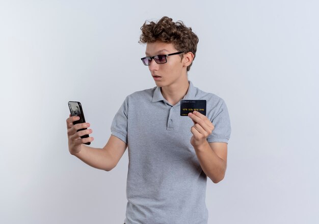 Młody człowiek w szarej koszulce polo patrząc na ekran swojego smartfona przedstawiający zdezorientowaną kartę kredytową stojącą na białej ścianie