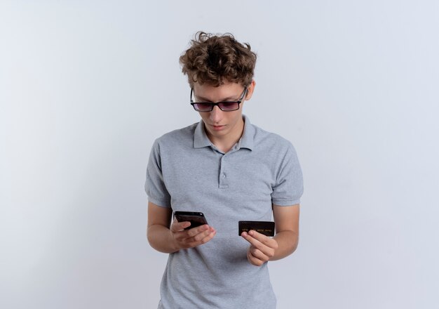 Młody człowiek w szarej koszulce polo, patrząc na ekran swojego smartfona trzymając kartę kredytową mylić stojąc na białej ścianie