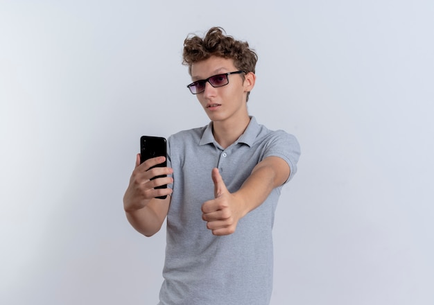 Młody Człowiek W Szarej Koszulce Polo Trzymając Smartfon Pokazując Kciuk Do Góry Stojąc Na Białej ścianie