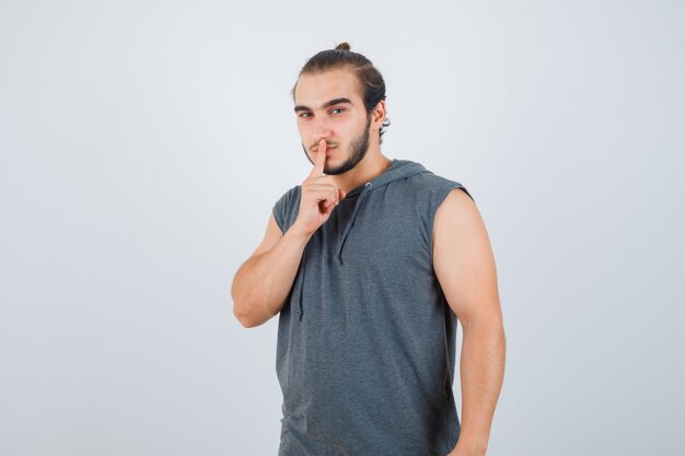 Młody człowiek w t-shirt z kapturem pokazujący gest ciszy i wyglądający poważnie, widok z przodu.