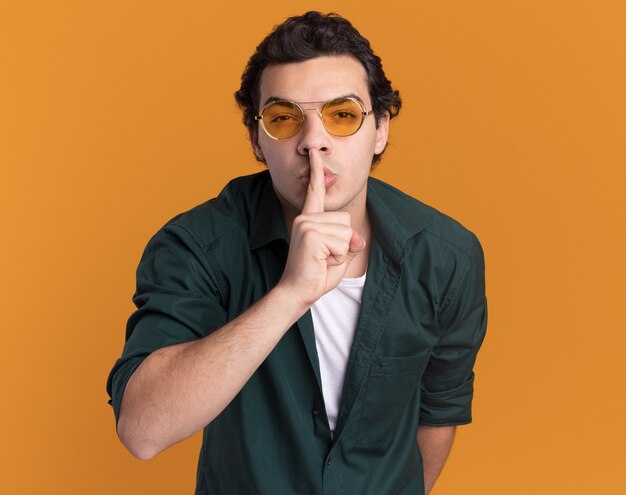 Młody człowiek w zielonej koszuli w okularach patrząc na przód robi gest ciszy z palcem na ustach stojąc na pomarańczowej ścianie