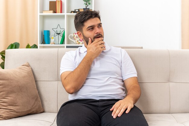 Młody człowiek w zwykłych ubraniach, patrząc na bok z ręką na brodzie, zdziwiony siedzi na kanapie w jasnym salonie