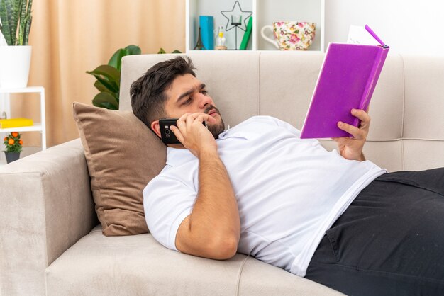 Młody człowiek w zwykłych ubraniach, trzymając książkę, czytając książkę i rozmawiając przez telefon komórkowy z poważną twarzą, spędzając weekend w domu leżąc na kanapie w jasnym salonie