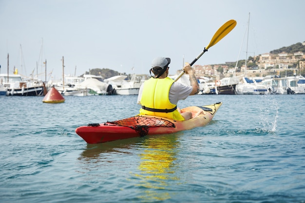 Młody człowiek wiosłujący na czerwonym kajaku na morzu w pobliżu statków i jachtów. Turysta robiący chlapanie wiosłem kajaka.