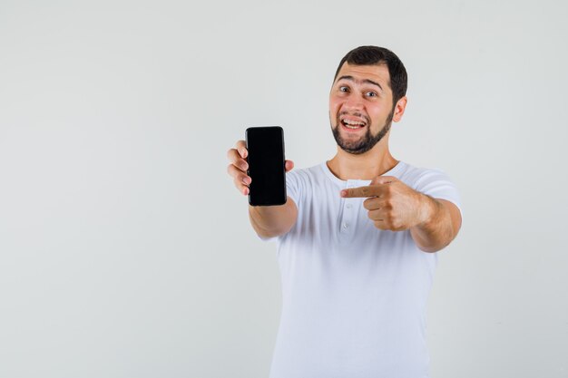 Młody człowiek, wskazując na telefon komórkowy w białej koszulce i patrząc szczęśliwy, przedni widok. miejsce na tekst