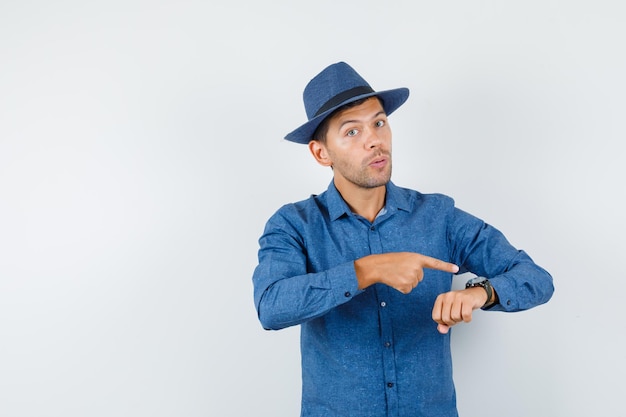 Młody człowiek wskazując na zegarek na nadgarstku w niebieskiej koszuli, kapeluszu i patrząc ostrożnie. przedni widok.