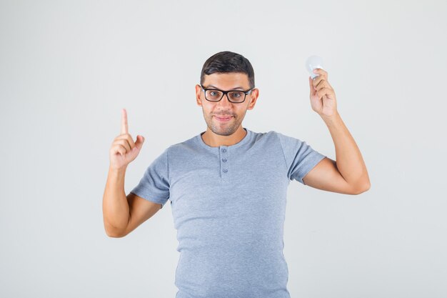 Młody człowiek wskazując palcem w górę i trzymając żarówkę w szarej koszulce, widok z przodu okulary.