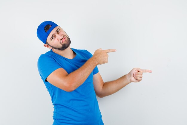 Młody człowiek wskazujący na bok w niebieskiej koszulce i czapce i wyglądający na niezadowolonego