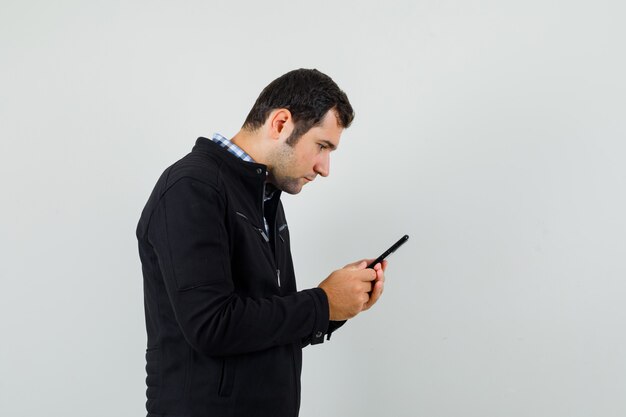 Młody człowiek za pomocą telefonu komórkowego w koszuli, kurtce i patrząc zajęty.