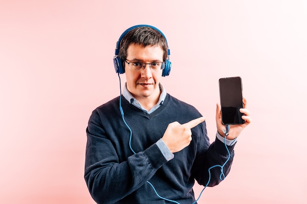 Młody dorosły mężczyzna trzydzieści pięć lat w koszuli i niebieskim swetrze z dekoltem w serek z różowym tłem, w okularach, słuchawkach muzycznych i smartfonie, pokazując smartfon i wskazując palcem
