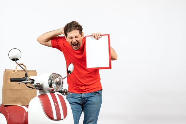 Młody emocjonalny nerwowy dostawa facet w czerwonym mundurze stojącym w pobliżu skutera pokazujący dokument na białej ścianie