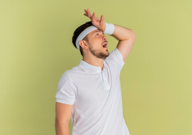 Młody fitness mężczyzna w białej koszuli z pałąkiem na głowę patrząc na bok mylić z ręką na głowie stojącej na oliwkowym tle