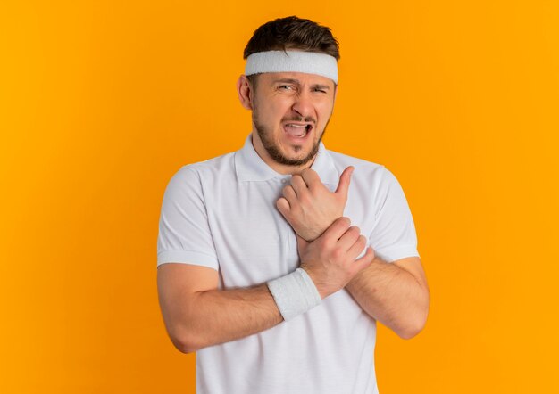 Młody fitness mężczyzna w białej koszuli z pałąkiem na głowę patrząc z przodu dotykając nadgarstka mając ból stojąc na pomarańczowej ścianie