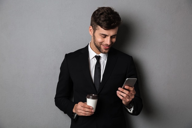 Młody handsom mężczyzna sprawdza wiadomość na smartphone w formalnej odzieży podczas gdy trzymający wynos kawę
