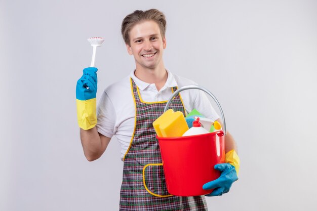 Młody hansdome mężczyzna ubrany w fartuch i gumowe rękawiczki, trzymając wiadro z narzędziami do czyszczenia i szorowania