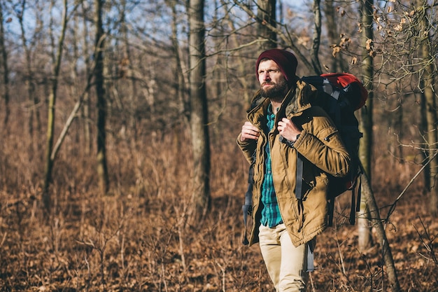 Młody hipster mężczyzna podróżujący z plecakiem w jesiennym lesie, ubrany w ciepłą kurtkę i kapelusz