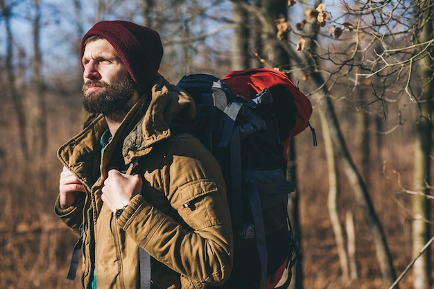 Młody hipster mężczyzna podróżujący z plecakiem w jesiennym lesie, ubrany w ciepłą kurtkę i kapelusz