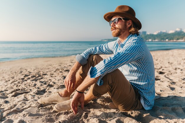 Młody hipster mężczyzna siedzi na plaży nad morzem na letnie wakacje, strój w stylu boho, ubrany w koszulę