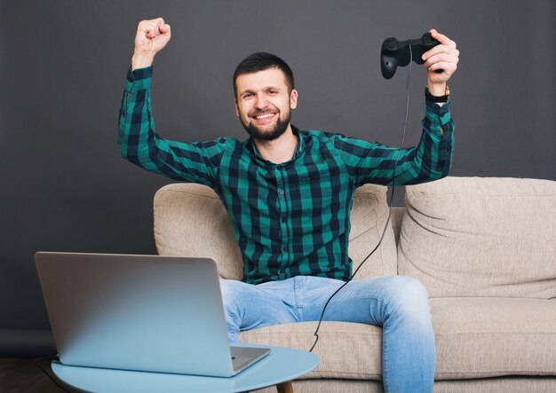 Młody hipster przystojny brodaty mężczyzna siedzi na kanapie w domu, grając w gry wideo na notebooku, trzymając joystick, zielona koszula w kratkę, szczęśliwy, uśmiechnięty, zabawa, rozrywka, świętuje zwycięstwo, ręce w górę