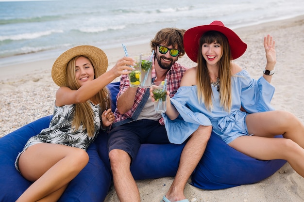 Bezpłatne zdjęcie młody hipster towarzystwo przyjaciół na wakacjach w kawiarni na plaży, pijąc koktajl mojito, szczęśliwy pozytywny, letni styl, uśmiechnięty szczęśliwy, dwie kobiety i mężczyzna bawią się razem, rozmawiają, flirt, romans, trzy