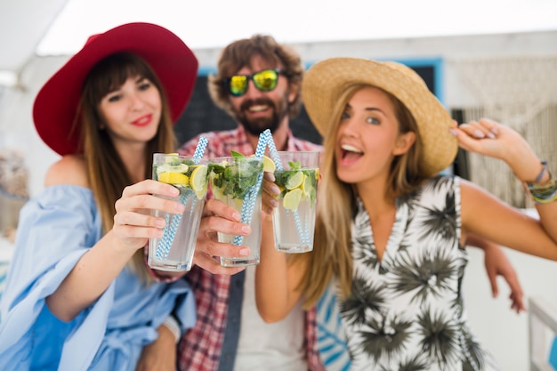 Bezpłatne zdjęcie młody hipster towarzystwo przyjaciół na wakacjach w letniej kawiarni, picie koktajli mojito, szczęśliwy pozytywny styl, uśmiechnięty szczęśliwy, dwie kobiety i mężczyzna bawią się razem, rozmawiają, flirt, romans, trzy