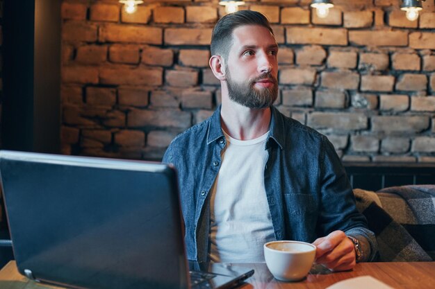 Młody hipster w barze z cappuccino, młody człowiek pije kawę w kawiarni w mieście w czasie obiadu i pracuje na laptopie