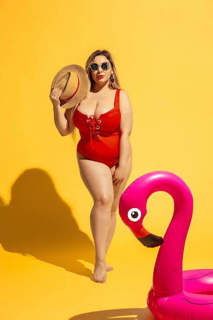 Młody kaukaski plus rozmiar modelki przygotowuje wakacje na żółtej ścianie. Kobieta w czerwonym stroju kąpielowym i kapeluszu pozuje z pływaniem.