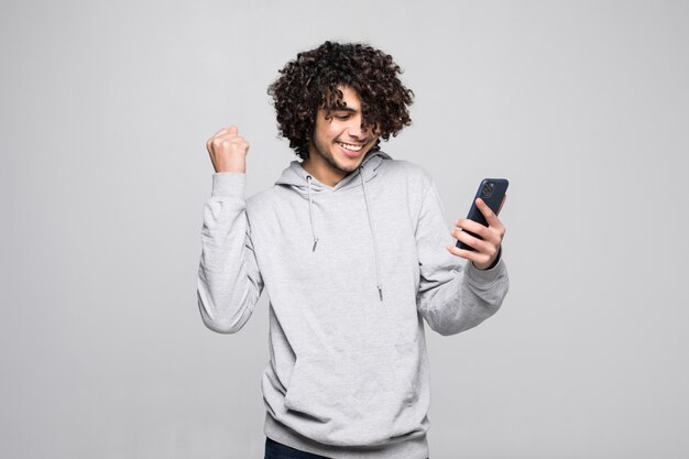 Młody kędzierzawy mężczyzna bawić się z telefonem patrzeje szczęśliwy i pięścią po wygranej, odizolowywający na białej ścianie.