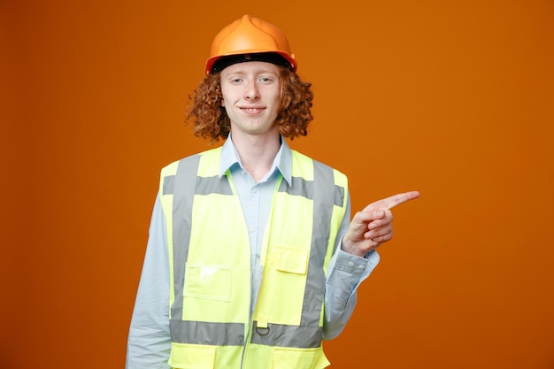 Młody konstruktor w mundurze budowlanym i kasku ochronnym, patrząc na kamerę, uśmiechając się pewnie, wskazując palcem wskazującym w bok, stojąc na pomarańczowym tle