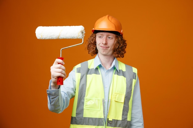 Bezpłatne zdjęcie młody konstruktor w mundurze budowlanym i kasku ochronnym, trzymający wałek do malowania, patrząc na niego szczęśliwy i zadowolony, uśmiechnięty, stojący na pomarańczowym tle