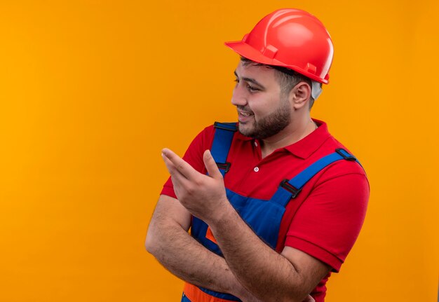 Bezpłatne zdjęcie młody konstruktor w mundurze konstrukcyjnym i kasku ochronnym patrząc na bok podnosząc rękę, zadając pytanie