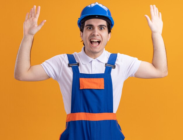 Młody konstruktor w mundurze konstrukcyjnym i kasku ochronnym patrząc na przód zdumiony i zaskoczony podnosząc dłonie stojąc nad pomarańczową ścianą