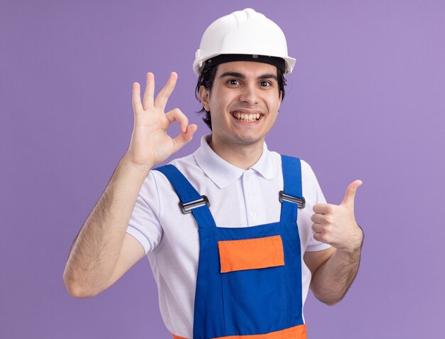 Młody konstruktor w mundurze konstrukcyjnym i kasku ochronnym patrząc z przodu szczęśliwy i pozytywny, pokazując znak ok i kciuki do góry stojąc nad fioletową ścianą