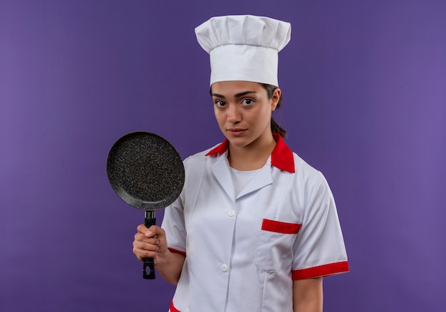 Bezpłatne zdjęcie młody kucharz kaukaski dziewczyna w mundurze szefa kuchni trzyma patelnię i patrzy na aparat na białym tle na fioletowym tle z miejsca na kopię